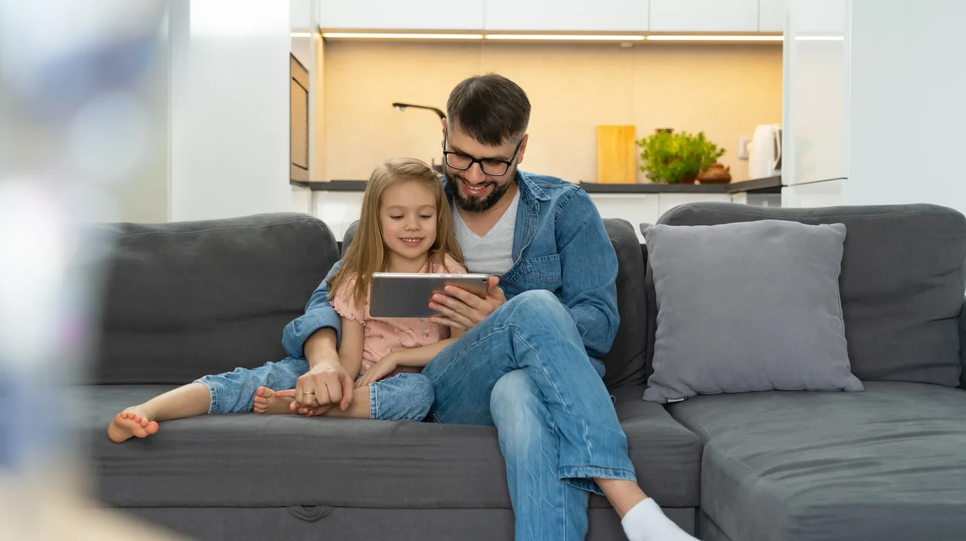 Een vader en zijn dochter kijken samen op een tablet op de bank thuis